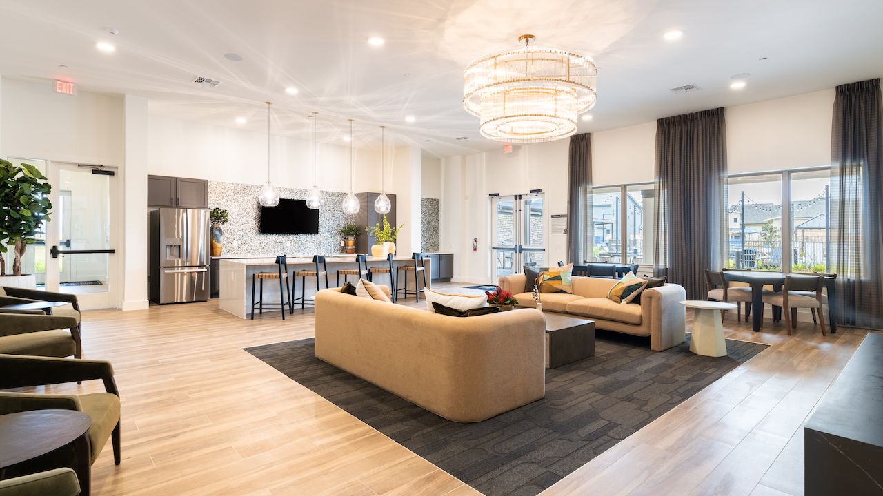 Modern interior of a community clubhouse featuring a seating area with beige sofas, a kitchen with bar seating, and a large chandelier above, with bright natural lighting